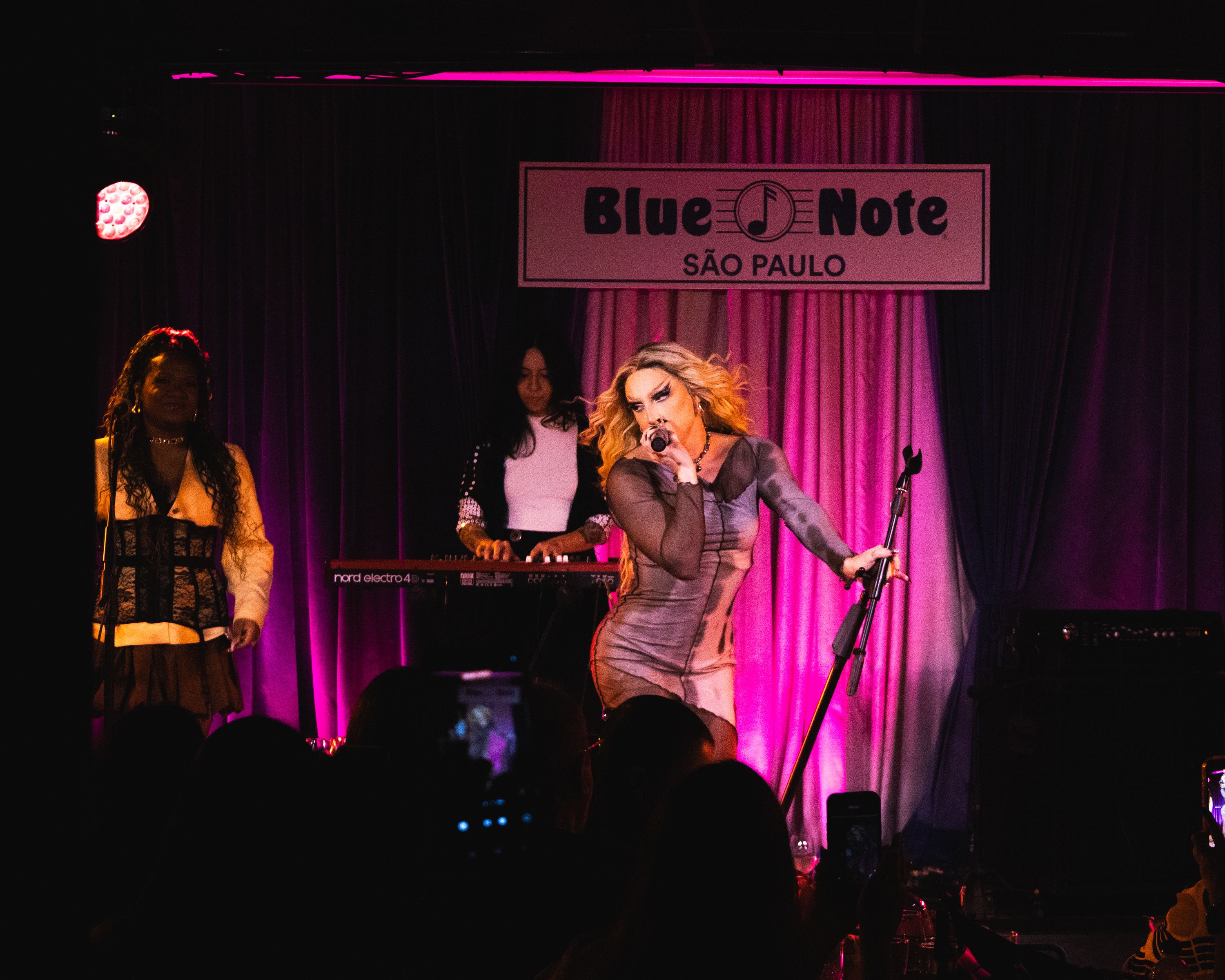 Diego Martins cantando no palco do Blue Note São Paulo, vestindo um vestido justo de mangas longas cinza, cabelo loiro ao vento, luzes rosas ao fundo e uma placa com o nome do local acima
